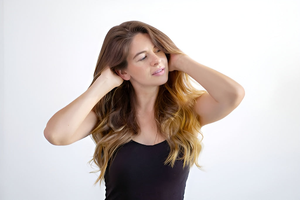 A young woman with long, brown, wavy hair, massaging her scalp with her hands.